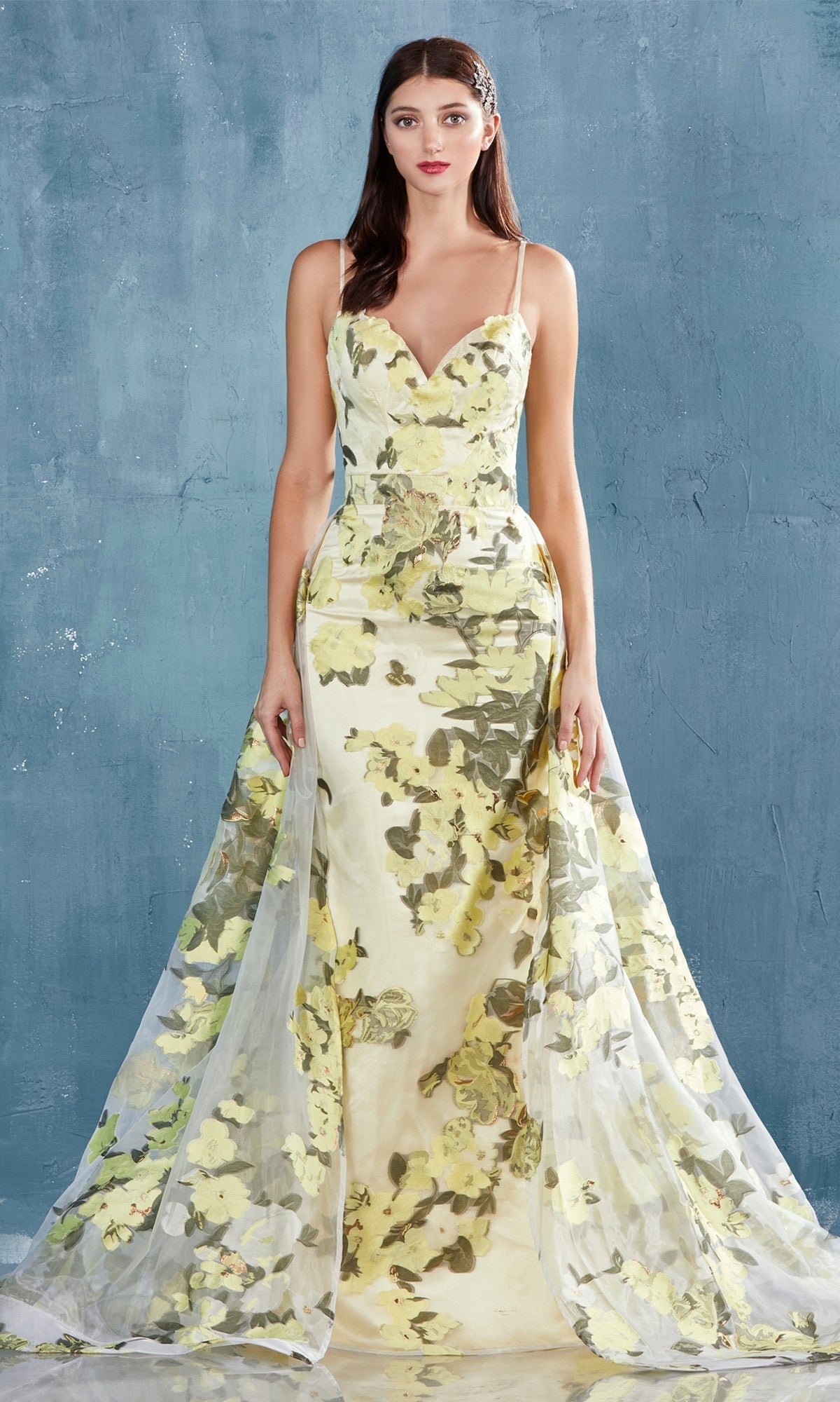 A woman stands against a textured blue wall wearing the Lemon Yellow Floral-Print Long Prom Dress A0770, a sleeveless, floor-length evening gown featuring a floral pattern and sheer overlay.